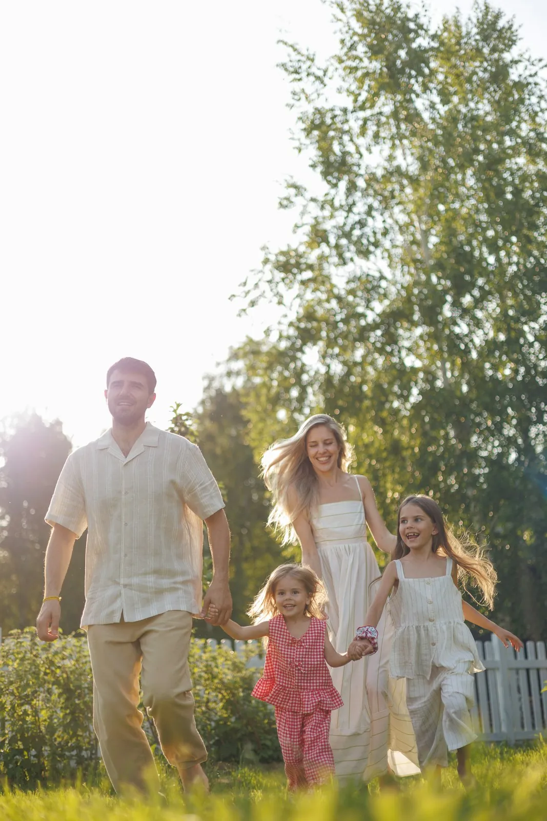 A father, mother and two young daughters run hand in hand across a sunlit lawn in front of a white picket fence.