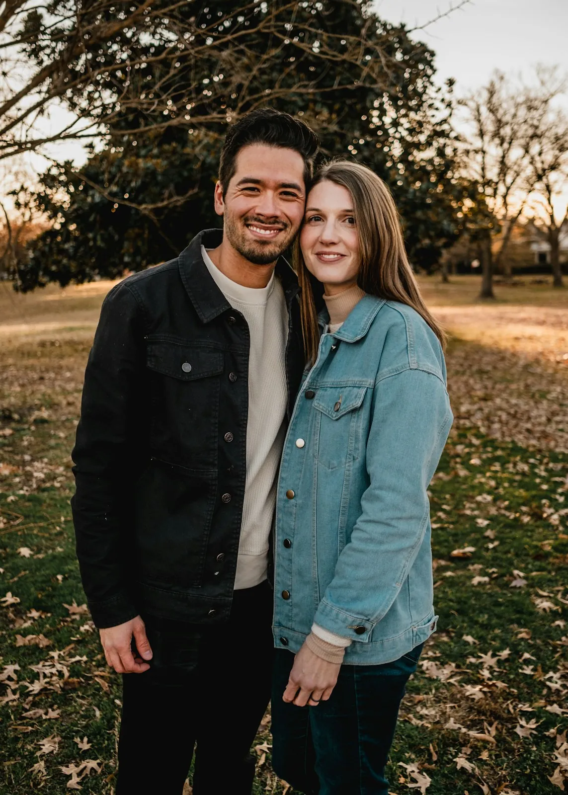 A smiling couple in denim jackets stand cheek to cheek in a park with fallen leaves and bare trees in late light.