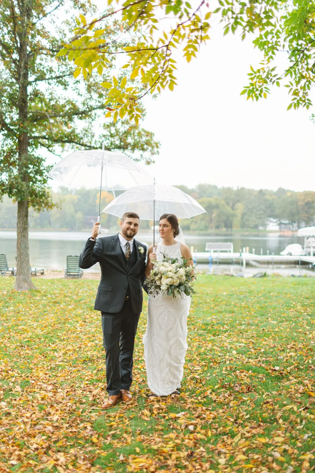 A bride holding a white bouquet and a groom in a dark suit stand under clear umbrellas on a leaf-covered lawn beside a lake.