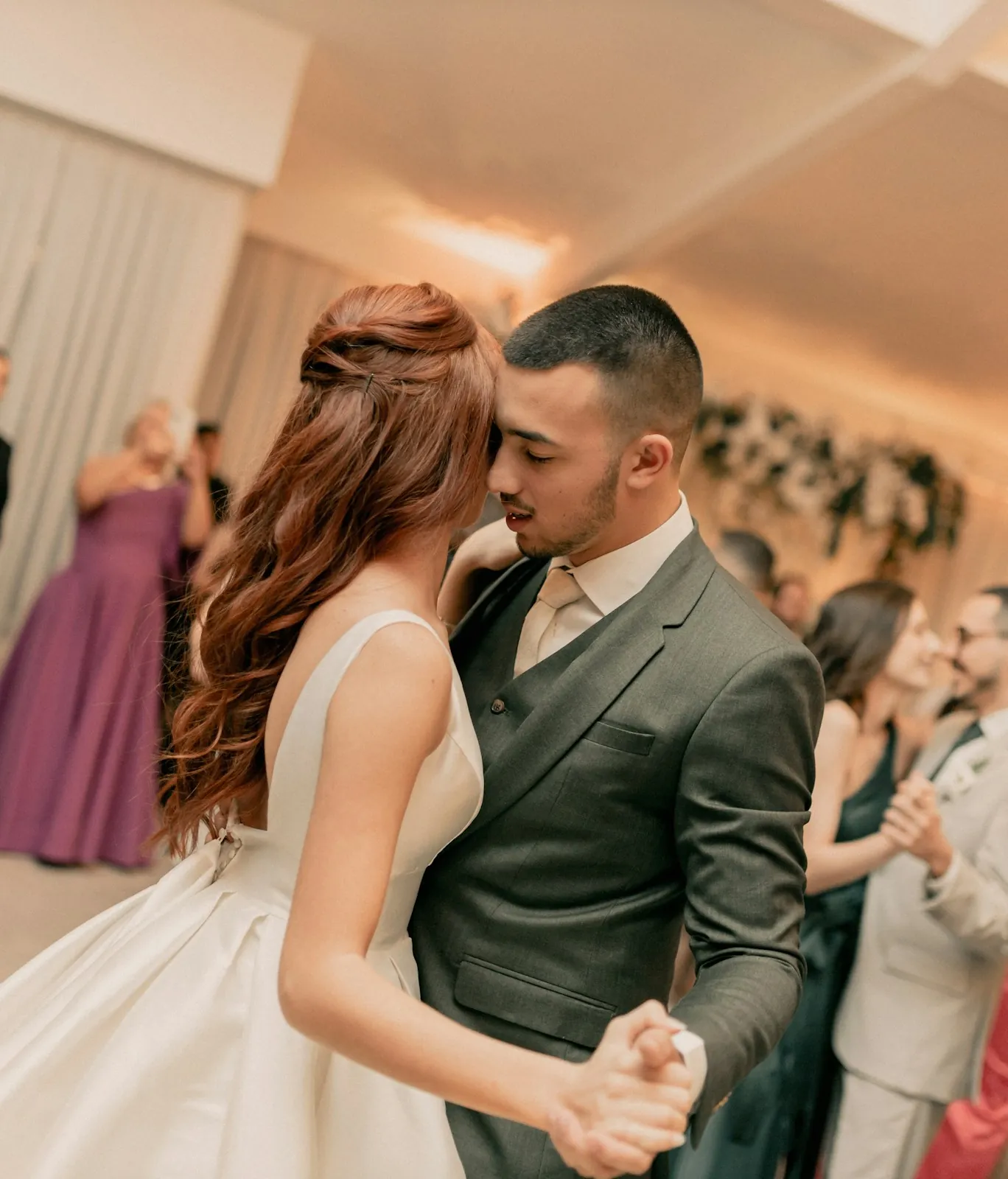A red-haired bride and a groom in a gray suit dance cheek to cheek while guests dance around them in a draped reception tent.