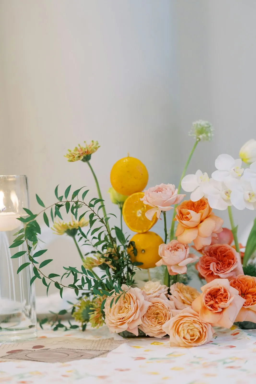 A low table centerpiece of peach garden roses, white orchids, greenery and halved oranges beside a glass candle holder.