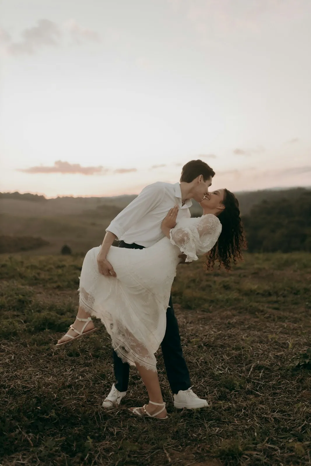 A man dips and kisses a woman in a white lace dress on a grassy hillside at dusk.