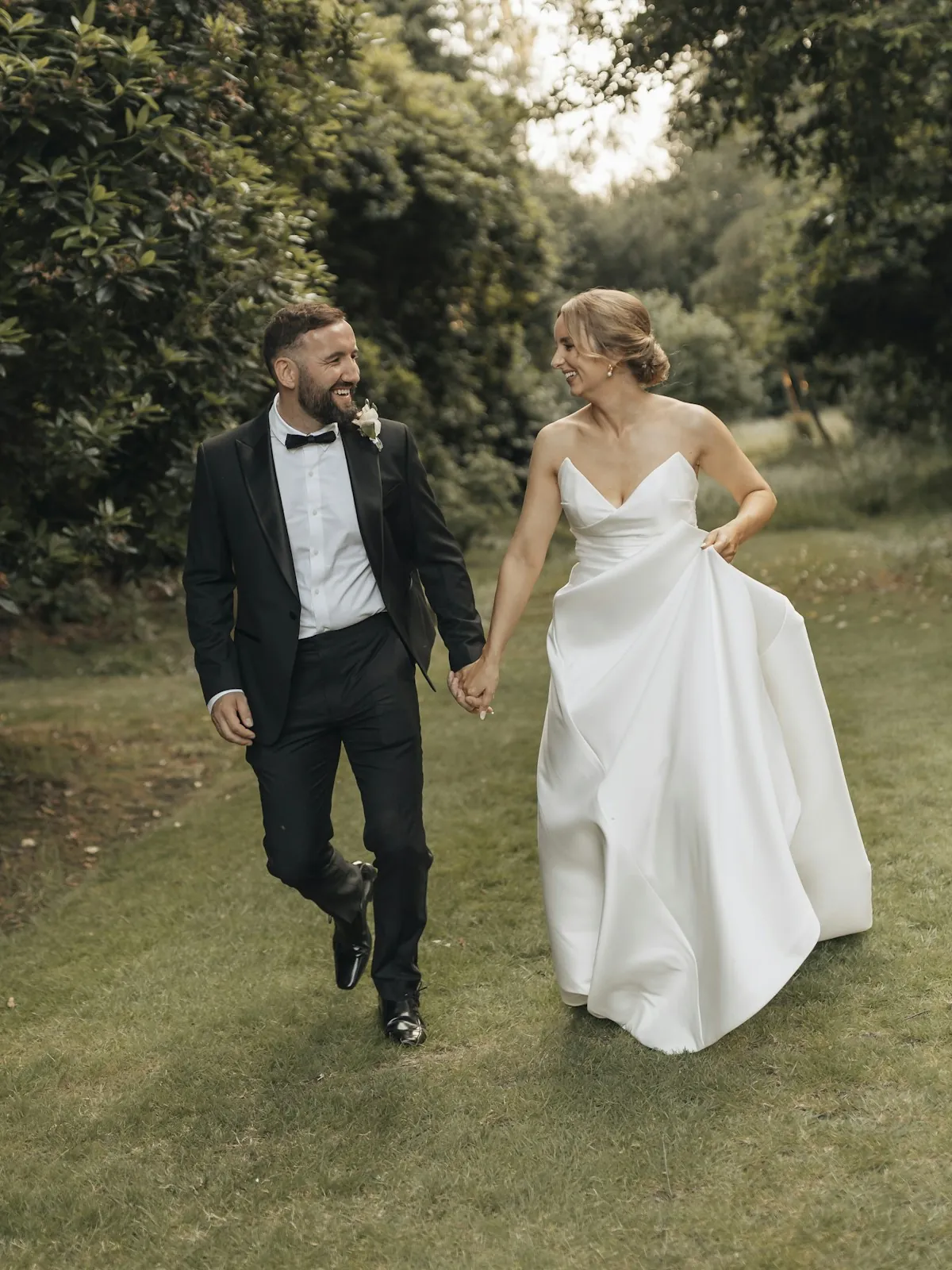 A bride in a strapless satin gown and a groom in a tuxedo walk hand in hand across a lawn between tall green shrubs, smiling at each other.