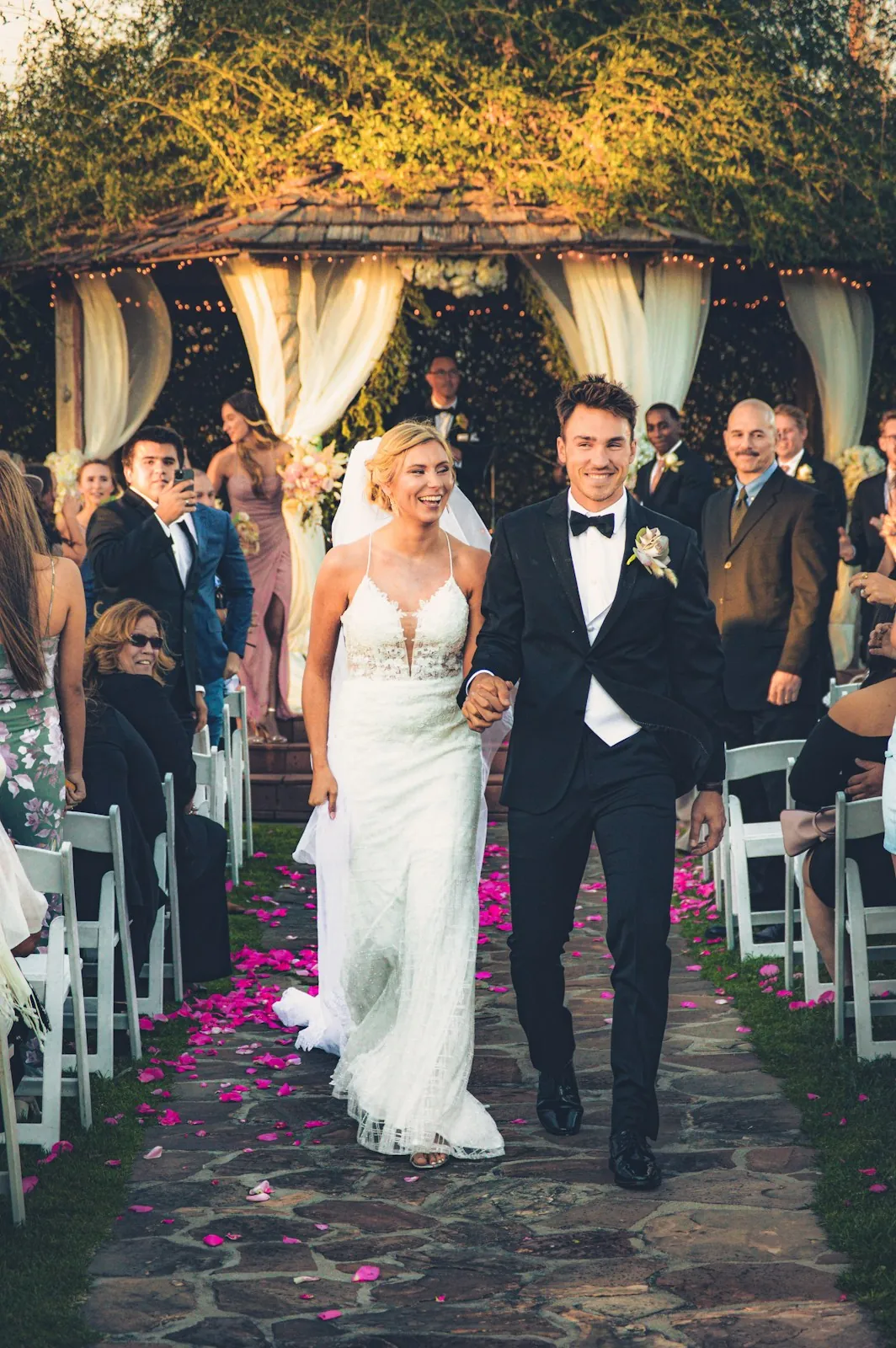 A laughing bride and groom walk back up a stone aisle scattered with pink petals as seated guests watch and take photos.