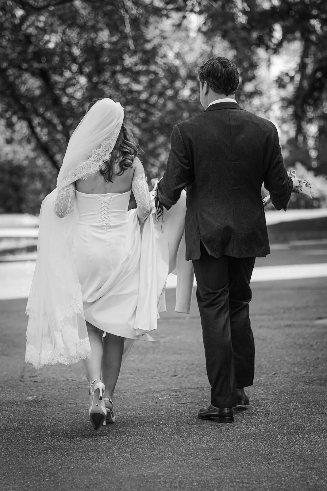 Black-and-white photo of a bride with a lace-edged veil and a groom in a dark suit walking away from the camera down a tree-lined path.