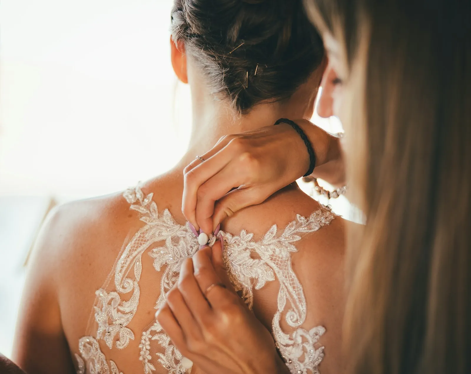 A woman fastens the small buttons on the lace back of a bride's gown in soft window light.