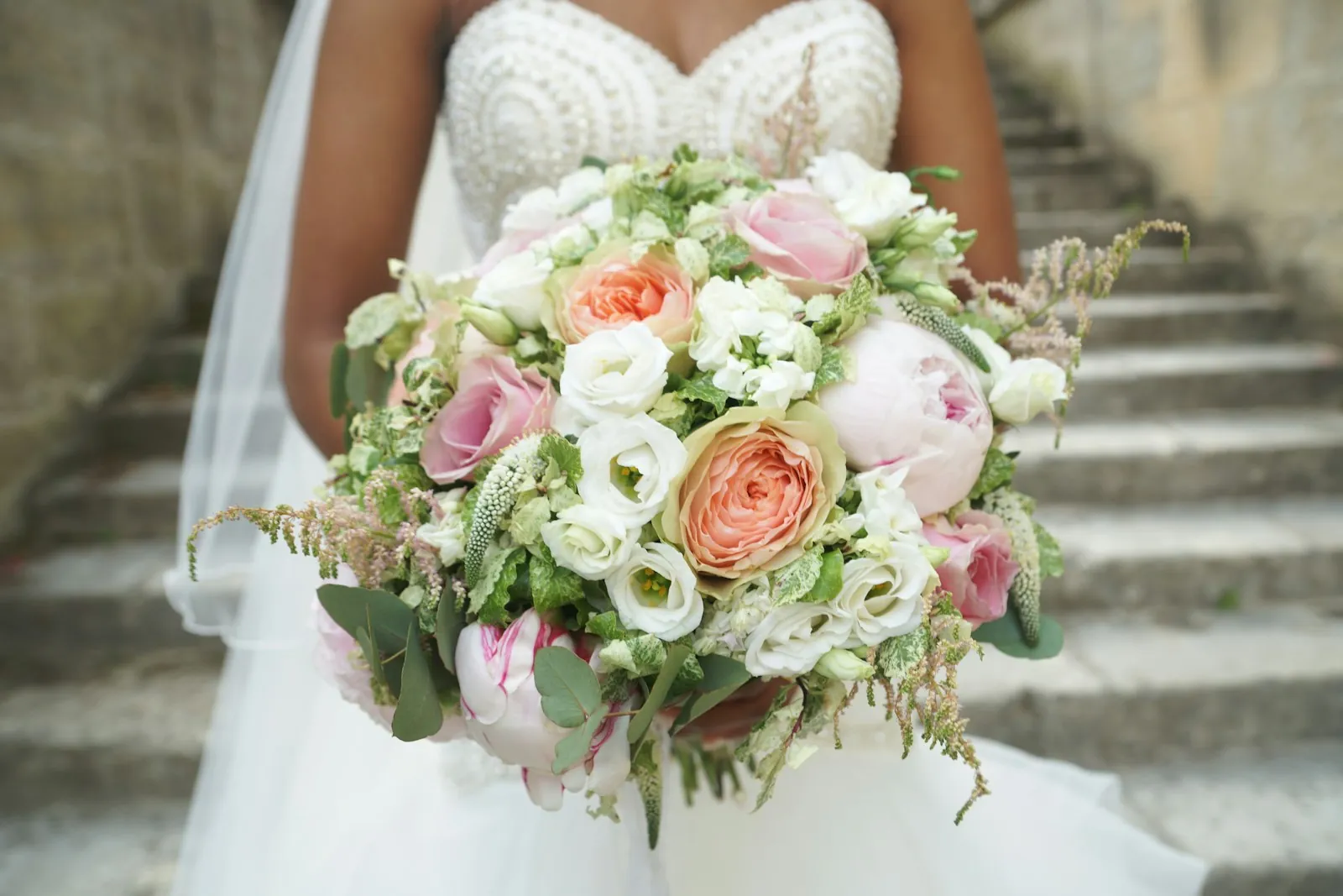 A bride in a beaded bodice holds a bouquet of pink peonies, peach garden roses and white lisianthus on stone steps.