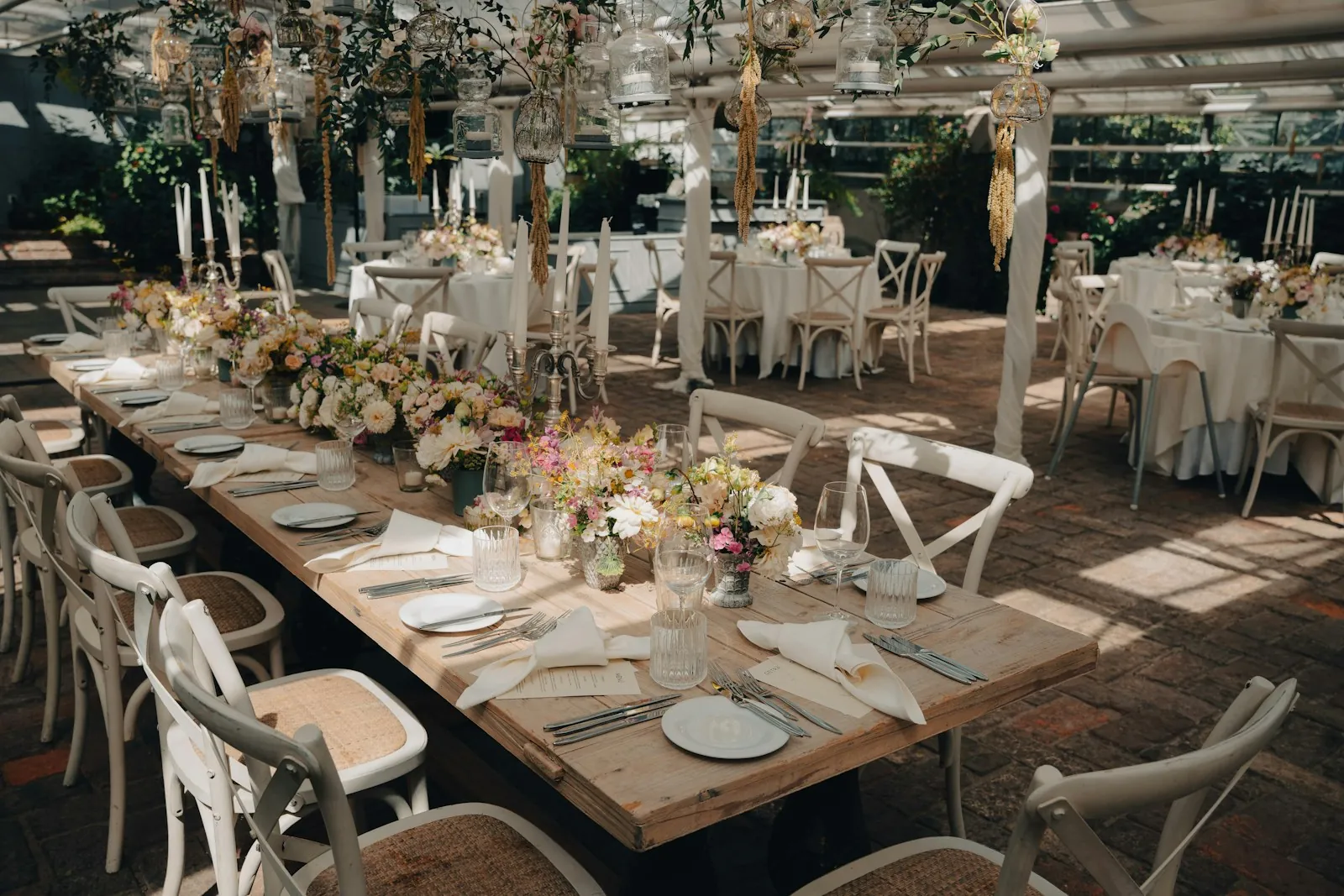 A long wooden table set with white plates, folded napkins and small vases of garden flowers inside a greenhouse reception space.