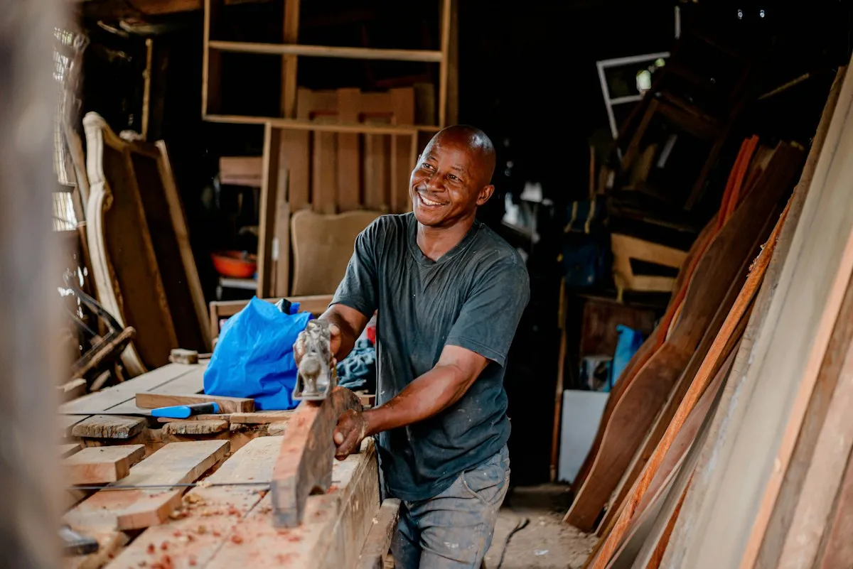 Carpenter smiling while planing a board in a wood-filled workshop