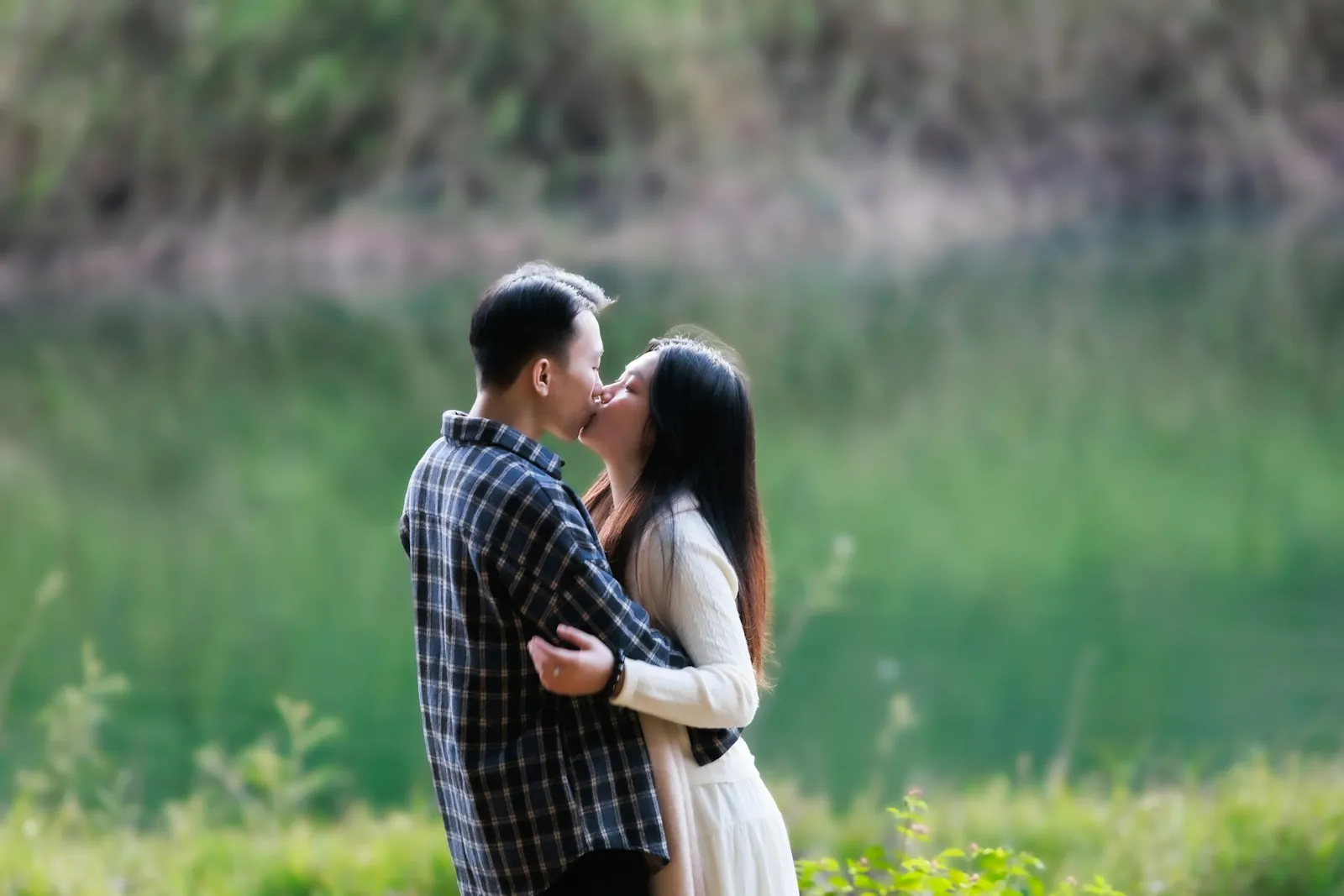 A couple in a plaid shirt and a white dress kiss beside a green pond with blurred grasses around them.