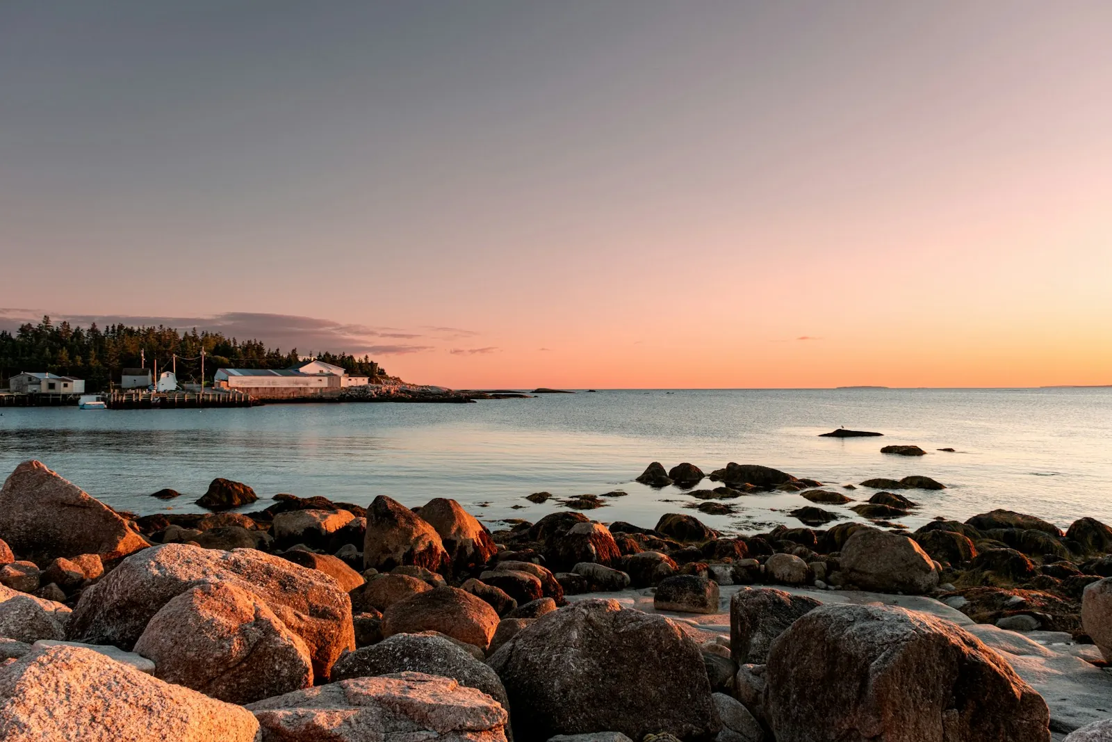 Granite boulders line a calm bay at dusk, with a small harbor and pine trees on the far shore under a pink sky.