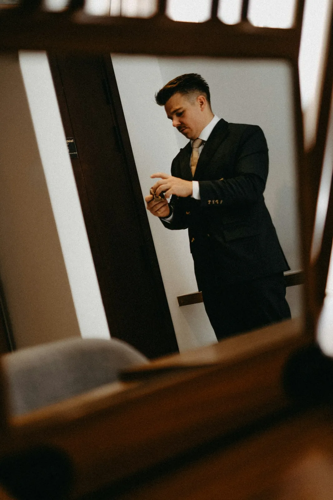 A groom in a navy suit adjusts his cufflinks, seen through the frame of a wooden mirror.