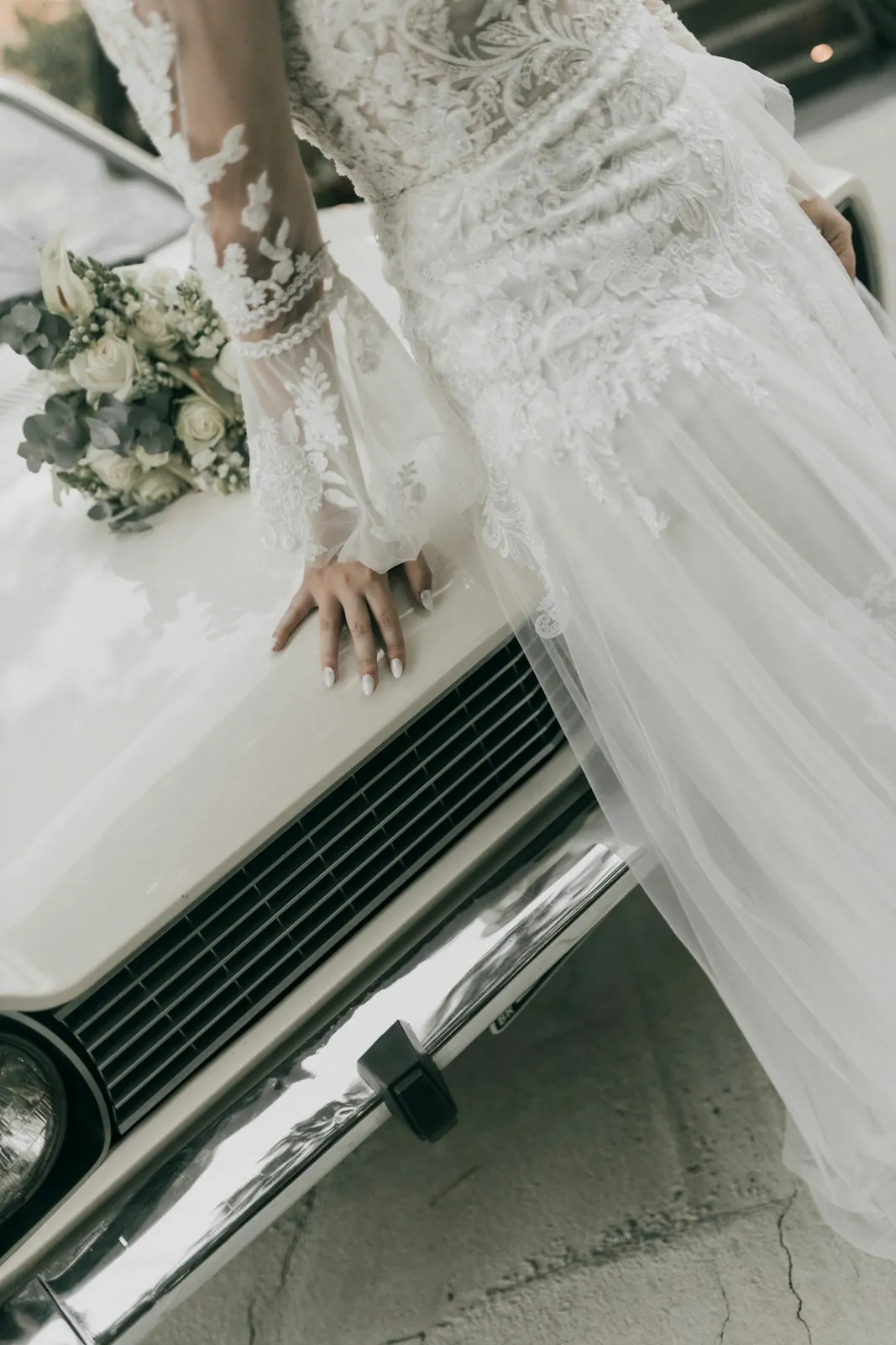 A bride in a lace gown rests her hand and bouquet on the hood of a white classic car with a chrome bumper.