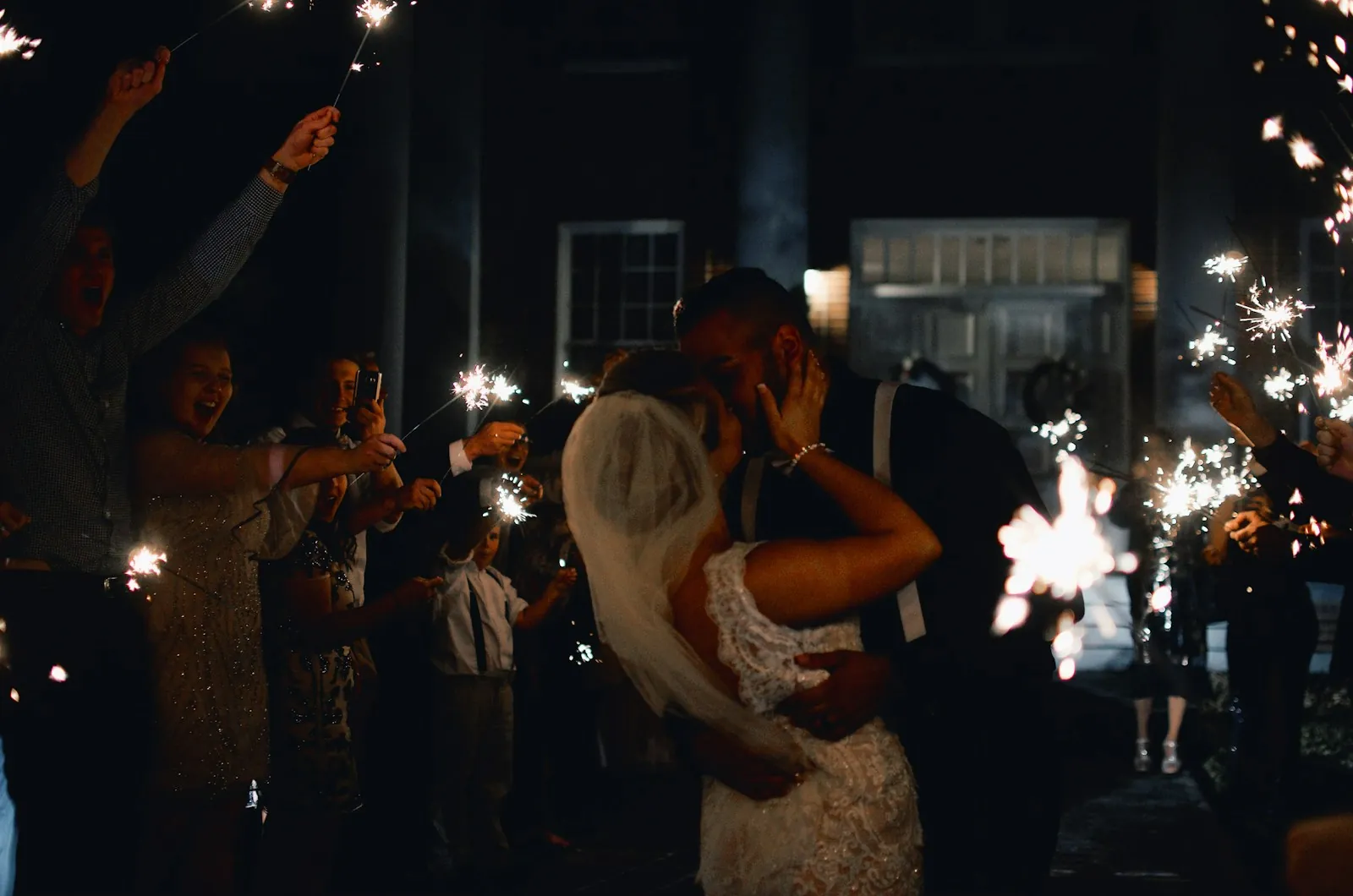 A bride and groom kiss at night while guests on both sides hold up lit sparklers and cheer.