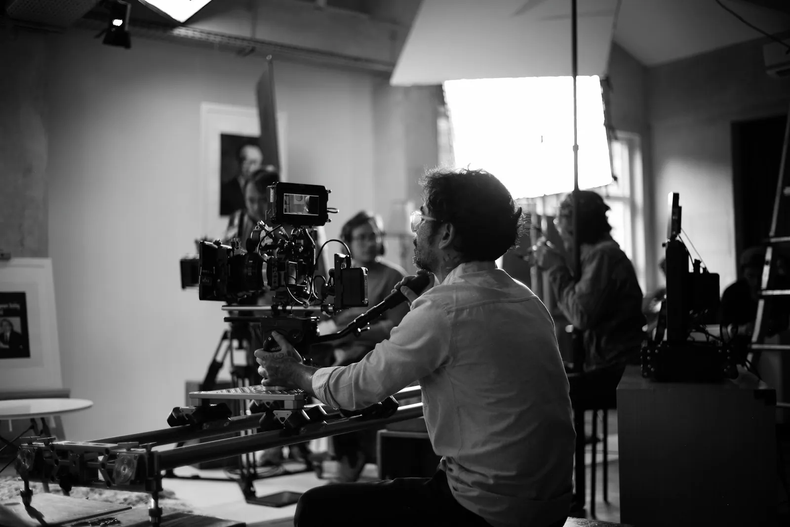 Black-and-white photo of a camera operator seated at a cinema camera on a slider while crew members work on a lit indoor set.