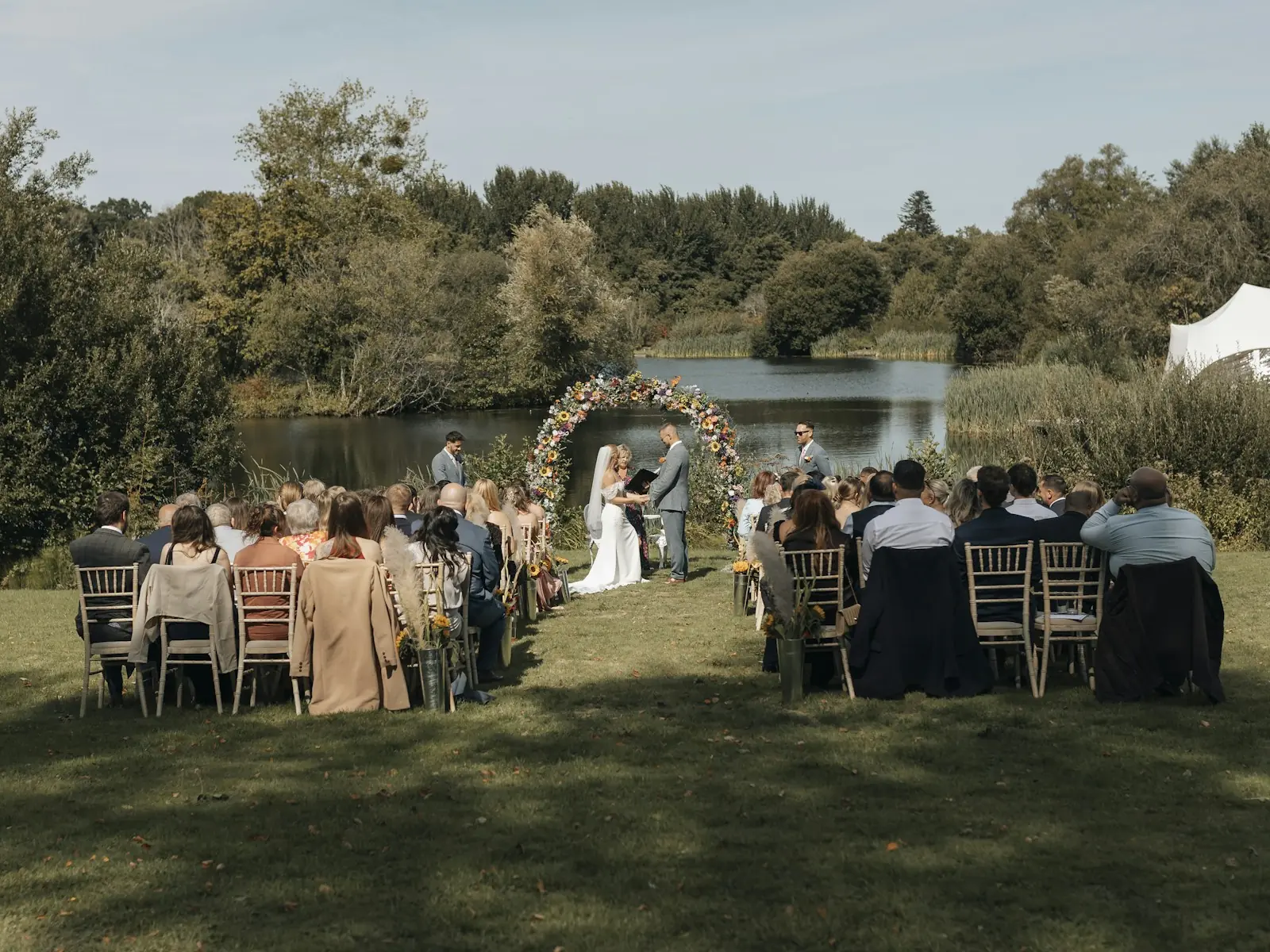Guests seated on wooden chairs watch a couple exchange vows under a floral arch beside a tree-lined lake.