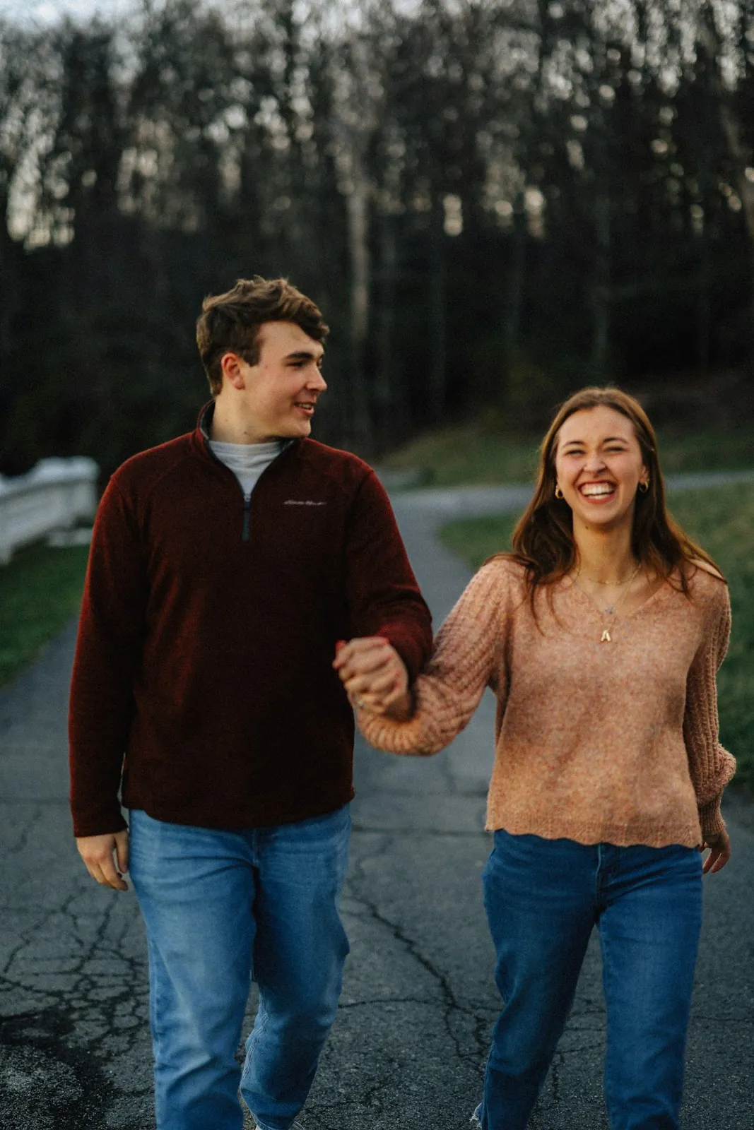 A laughing couple in sweaters and jeans walk hand in hand along a paved path at the edge of a forest.
