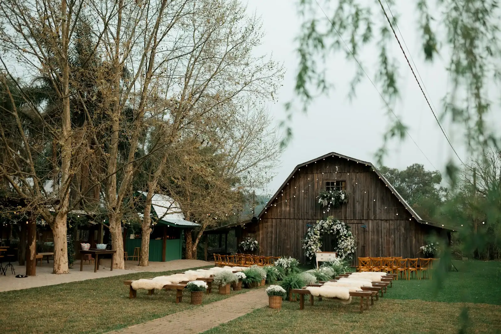 An old wooden barn strung with lights and a white floral wreath faces rows of benches draped with sheepskins on the lawn.