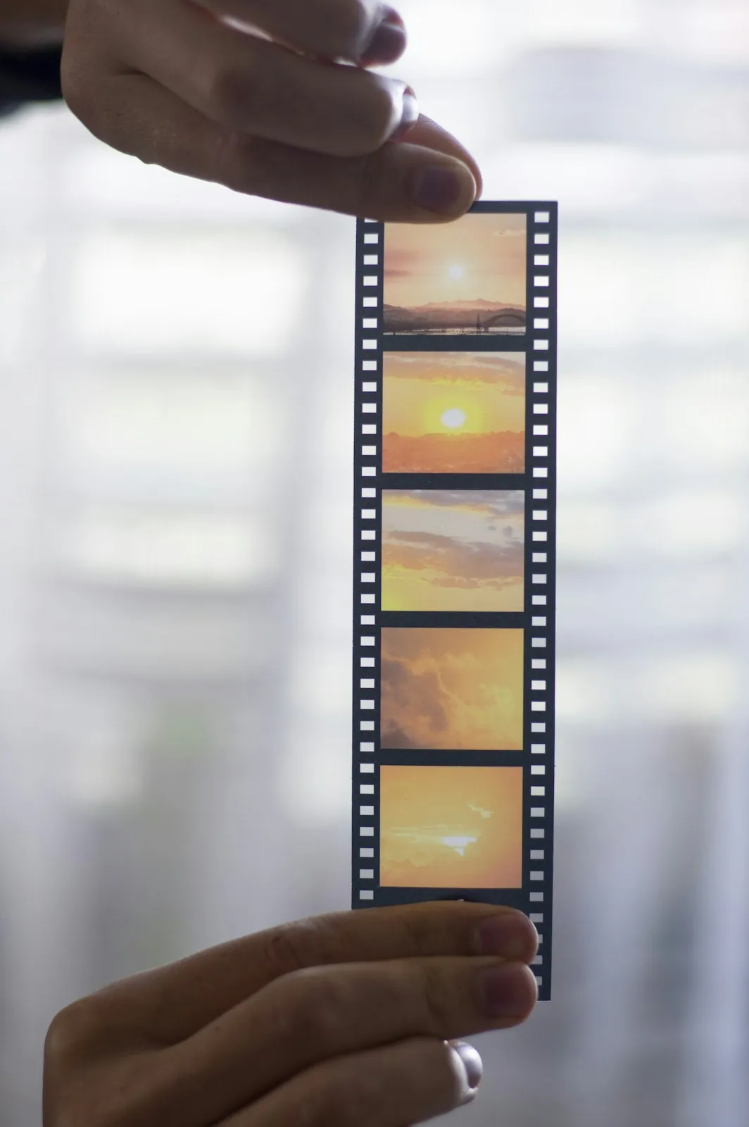 Two hands hold a strip of film frames showing an orange sunset up to a bright window.