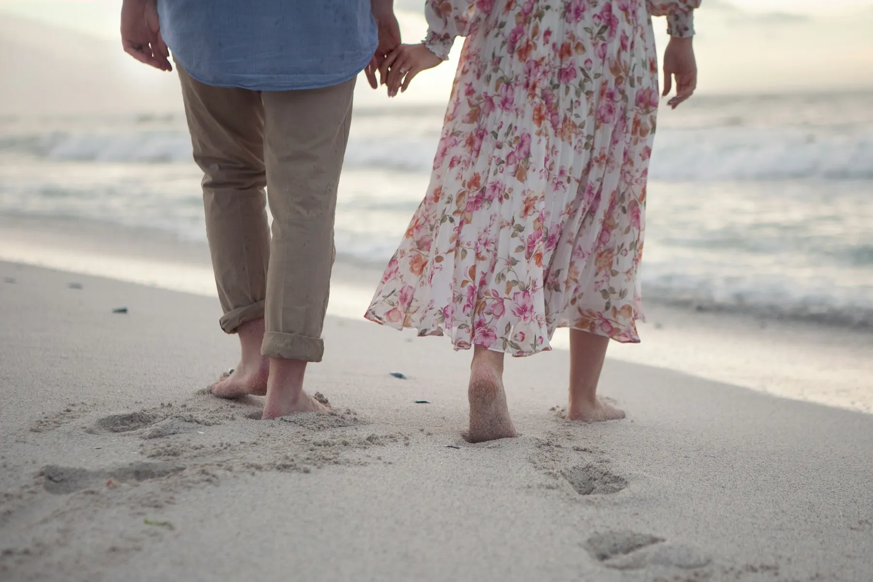 A barefoot couple holding hands, walking toward the surf at sunset