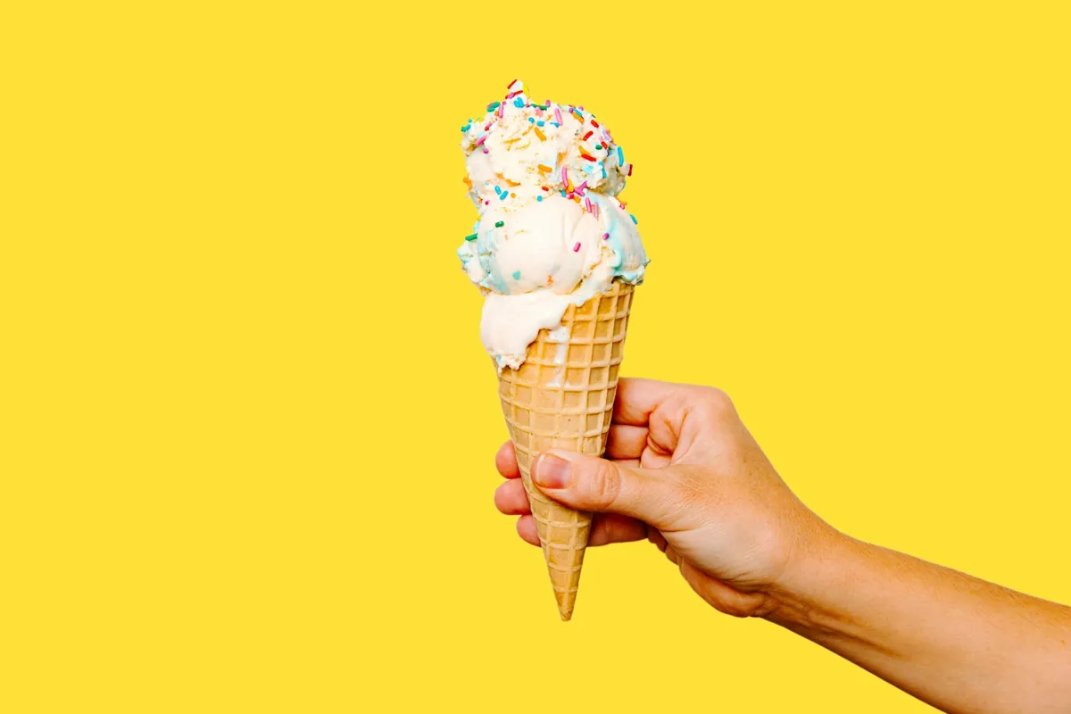 A hand holding up a vanilla cone covered in rainbow sprinkles against a bright yellow wall