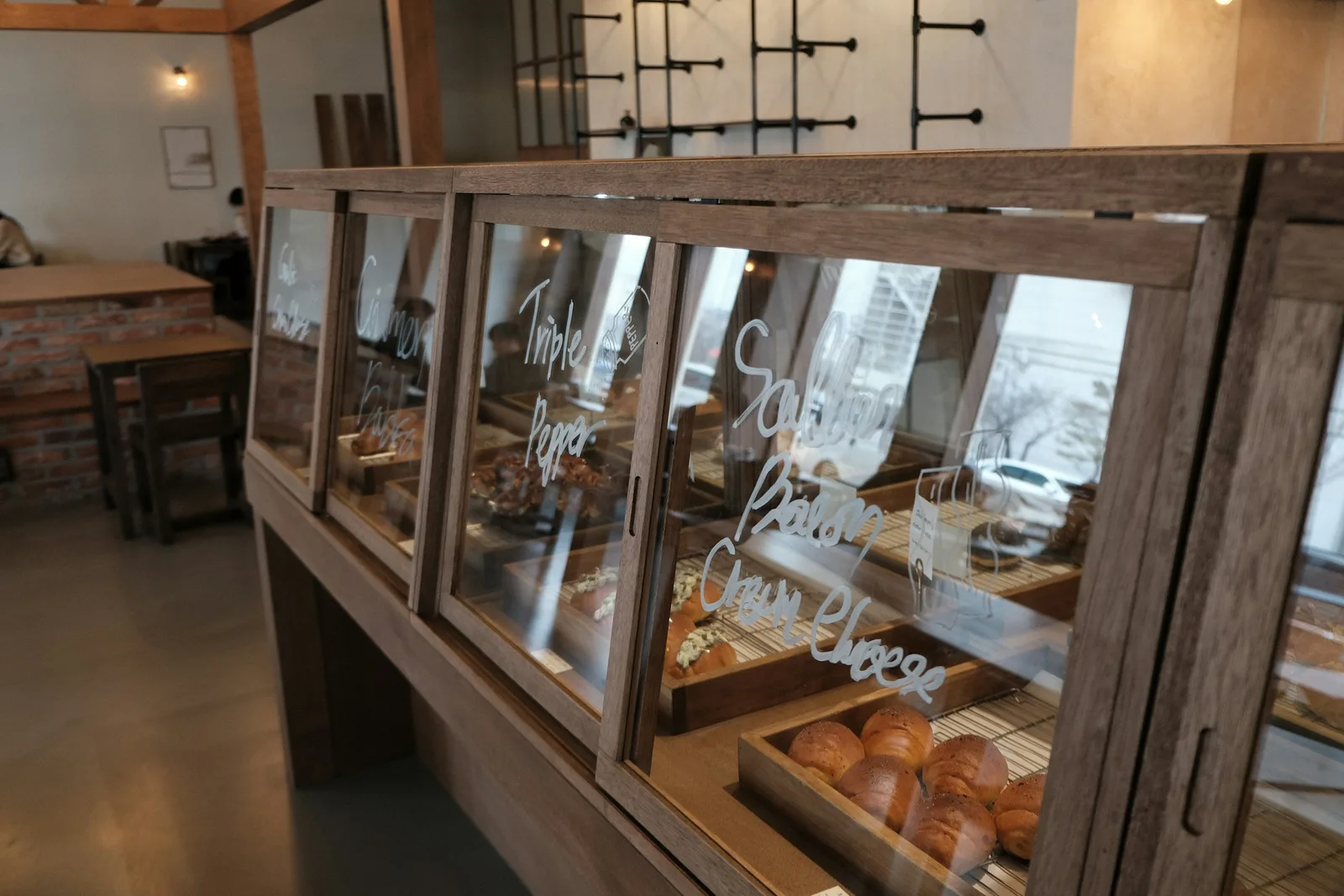A wood-framed glass bakery case with trays of rolls and pastries, café tables and a brick wall behind it