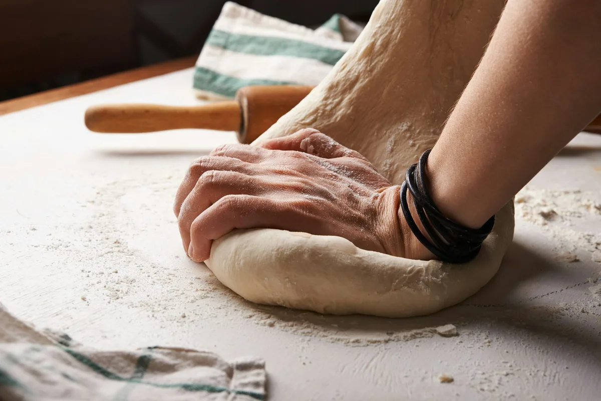 Hands kneading a round of dough on a floured bench beside a rolling pin